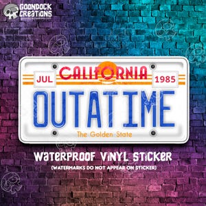 May include: A white California license plate with blue text that reads "OUTATIME" and "The Golden State". The plate has a red and orange sunset graphic and the text "JUL 1985". The plate is on a brick wall background.