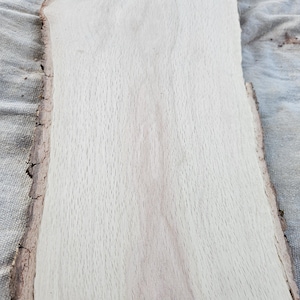 May include: A light-colored, unfinished wood slab with a natural edge. The wood grain is visible, and the slab has a rectangular shape with a rough, bark-like edge. The wood appears to be smooth and ready for use in a woodworking project.