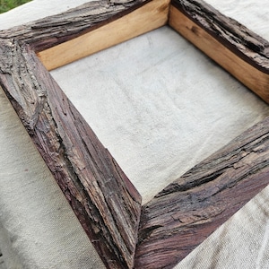 May include: Square wooden picture frame with a rustic, bark-like texture. The frame is made of natural wood with a dark brown color and visible grain. The frame's corners are joined at 90-degree angles.