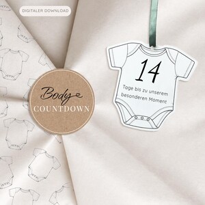 Può includere: Un cartoncino bianco a forma di body con un nastro verde, che riporta il numero 14 e il testo in tedesco 'Tage bis zu unserem besonderen Moment'. Accanto si trova un'etichetta rotonda marrone con 'Body COUNTDOWN' su uno sfondo a fantasia di body.