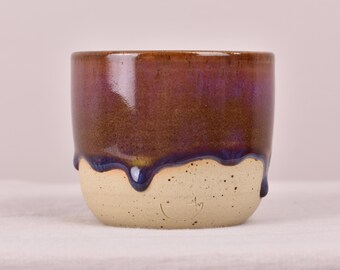 May include: A handmade ceramic cup with a unique glaze. The top half is a deep brown with purple hues, transitioning to a dark blue drip over a speckled, off-white base. The cup has a small, stamped design on the bottom.