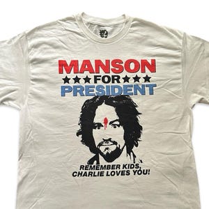 May include: Off-white t-shirt with the text "MANSON FOR PRESIDENT" in red and blue. Below the text is a black and white illustration of a man's face. The text "REMEMBER KIDS, CHARLIE LOVES YOU!" is printed below the illustration.