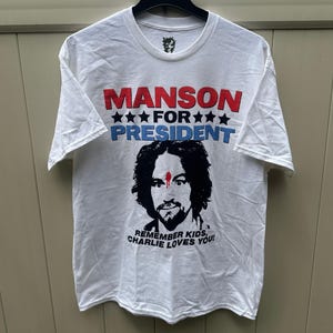 May include: White t-shirt with the text "MANSON FOR PRESIDENT" in red and blue, above a black and white portrait. The text "REMEMBER KIDS, CHARLIE LOVES YOU!" is below the portrait. The shirt is hanging on a black hanger.