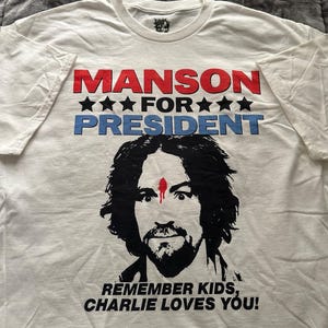 May include: White t-shirt with the text "MANSON FOR PRESIDENT" in red and blue, with black stars. Below is a black and white portrait of a man with a red mark on his forehead. The text "REMEMBER KIDS, CHARLIE LOVES YOU!" is at the bottom.