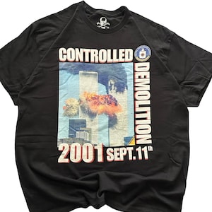 May include: A black short-sleeve t-shirt featuring a graphic print of the World Trade Center towers with an explosion. The text reads CONTROLLED DEMOLITION and 2001 SEPT. 11th, with a small circular emblem at the top right of the graphic design.