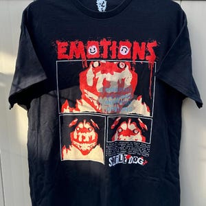 Può includere: T-shirt nera con un design grafico con la scritta "EMOTIONS" in rosso, con simboli di faccine sorridenti. Sotto, un'illustrazione a più pannelli di una creatura simile a un cane con una combinazione di colori rosso e bianco. È visibile anche il testo "SMILE DOG".