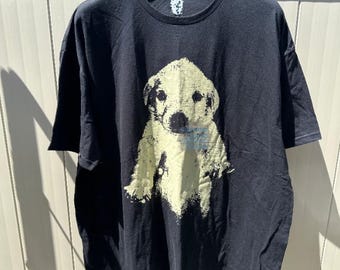 May include: Black t-shirt featuring a distressed, light yellow graphic of a puppy. The puppy has black eyes and nose. The shirt is hanging on a black hanger against a light-colored background.