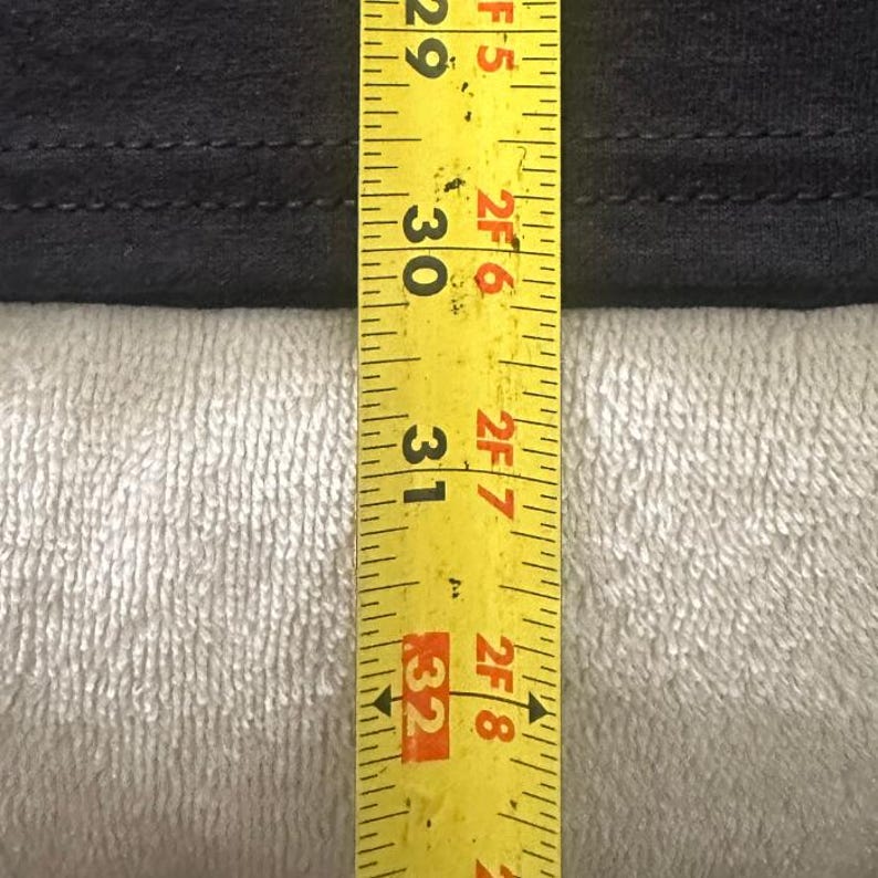 May include: A yellow measuring tape showing measurements in inches. The tape is next to a black fabric. The tape reads 29, 30, 31, and 32 inches. The fabric is on a white, textured surface.