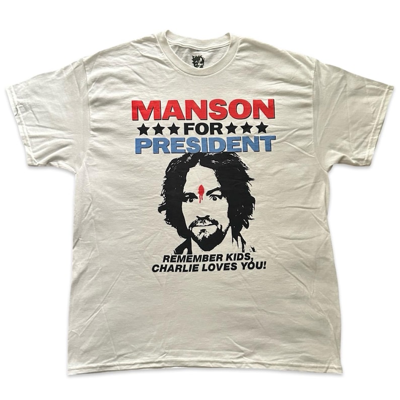 May include: Off-white t-shirt with the text "MANSON FOR PRESIDENT" in red and blue. Below the text is a black and white illustration of a man's face. The text "REMEMBER KIDS, CHARLIE LOVES YOU!" is printed below the illustration.