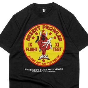 Puede incluir: Camiseta negra con un diseño gráfico circular que presenta una figura roja con capucha, estrellas y el texto "DESERT PROWLER". El texto adicional incluye "IX FLIGHT", "XI TEST" y "ALONE AND ON THE PROWL". Debajo, se lee "PENTAGON'S BLACK OPERATIONS".