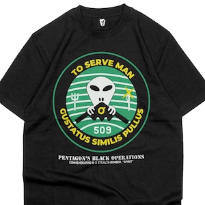 May include: Black t-shirt with a circular graphic featuring an alien head and the text "TO SERVE MAN". The graphic also includes the text "GUSTATUS SIMILIS PULLUS" and "509". Below the graphic is the text "PENTAGON'S BLACK OPERATIONS COMMEMORATING B-2 STEALTH BOMBER, 'SPIRIT'".