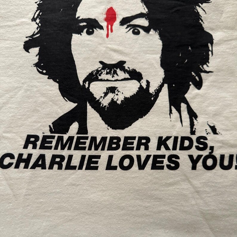 May include: White t-shirt featuring a black and white graphic of a man's face with a red dot on his forehead. The text "REMEMBER KIDS, CHARLIE LOVES YOU!" is printed in black below the graphic.