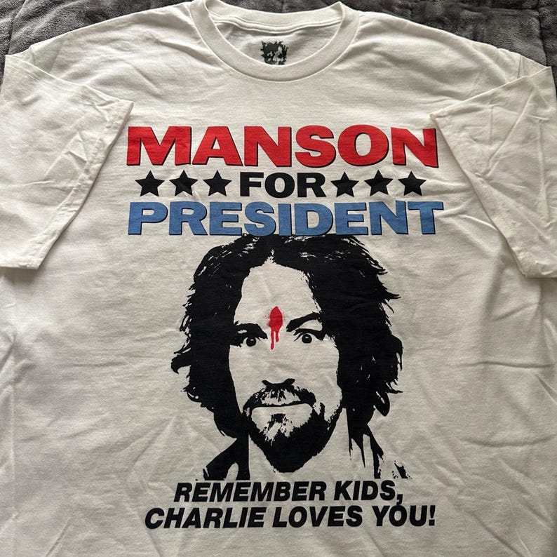 May include: White t-shirt with the text "MANSON FOR PRESIDENT" in red and blue, with black stars. Below is a black and white portrait of a man with a red mark on his forehead. The text "REMEMBER KIDS, CHARLIE LOVES YOU!" is at the bottom.