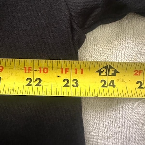 May include: A black garment is measured with a yellow tape measure. The tape measure shows measurements from 22 inches to 24 inches. The garment is on a white, textured surface.