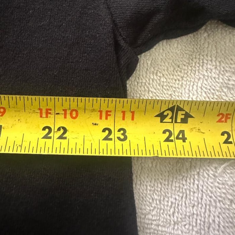 May include: A black garment is measured with a yellow tape measure. The tape measure shows measurements from 22 inches to 24 inches. The garment is on a white, textured surface.