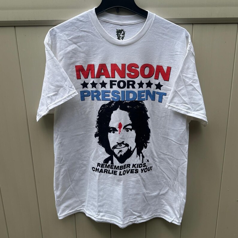 May include: White t-shirt with the text "MANSON FOR PRESIDENT" in red and blue, above a black and white portrait. The text "REMEMBER KIDS, CHARLIE LOVES YOU!" is below the portrait. The shirt is hanging on a black hanger.