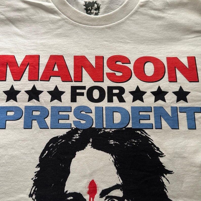 May include: White t-shirt with the text "MANSON FOR PRESIDENT" in red and blue, with black stars. The shirt features a black and white graphic of a person's face.
