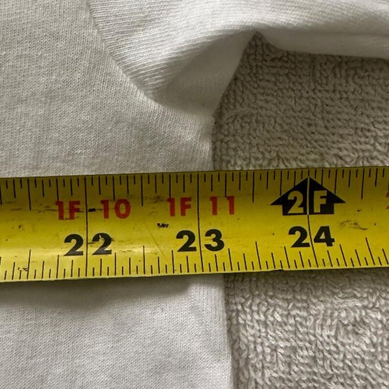 May include: Close-up of a white fabric with a yellow measuring tape. The tape shows measurements from 22 to 24 inches. The fabric is slightly wrinkled and appears to be a piece of clothing.