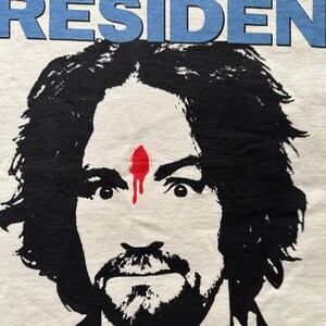 May include: White t-shirt featuring a black and white graphic portrait of a man with a beard and mustache. A red mark is on his forehead, with a dripping effect. The word "RESIDENT" is printed in blue above the portrait.