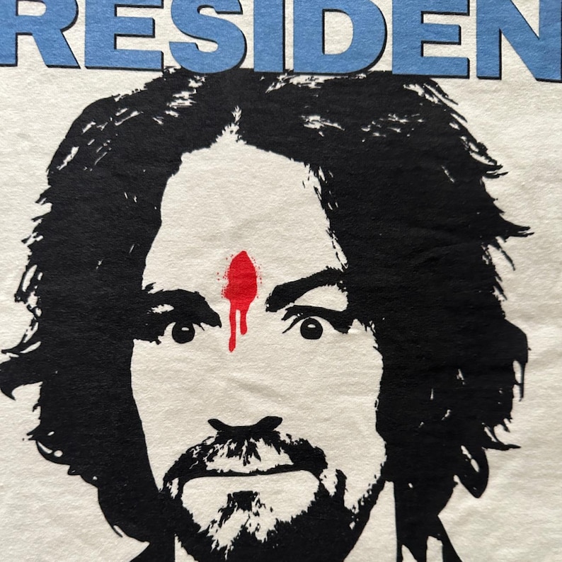 May include: White t-shirt featuring a black and white graphic portrait of a man with a beard and mustache. A red mark is on his forehead, with a dripping effect. The word "RESIDENT" is printed in blue above the portrait.