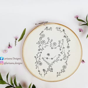 Può includere: Un telaio da ricamo con tessuto color crema, raffigurante un disegno floreale nero a forma di figura femminile. Il disegno è incorniciato da un cerchio di legno. I social media dell'artista sono visibili.
