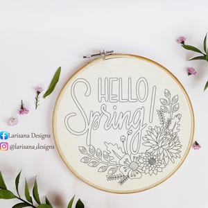 May include: An embroidery hoop with the words "HELLO Spring!" in black script and block letters. The design features floral and leaf outlines. The hoop is surrounded by small pink flowers and green leaves.