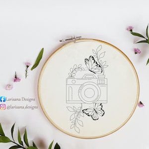 May include: A black and white line drawing of a camera, butterflies, and leaves is embroidered on a cream-colored fabric stretched within a wooden embroidery hoop. The design includes the text "Larisana Designs" and social media handles.