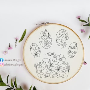 May include: A floral embroidery hoop featuring a paw print design with the word "love". The design is in black thread on a cream-colored fabric. The hoop is surrounded by small pink flowers and green leaves.