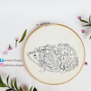 Op de afbeelding: Een handgeborduurde borduurring met een zwarte lijntekening van een egel gevuld met bladeren en bloemmotieven. Het ontwerp staat op een crèmekleurige stof, geplaatst in een houten ring. De tekst "Larisana Designs" is zichtbaar.