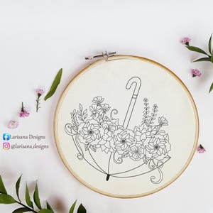May include: A cream-coloured fabric stretched in a wooden embroidery hoop, featuring a black line drawing of an umbrella overflowing with flowers. The image includes the text "Larisana Designs" and their social media handles.
