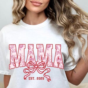 May include: White t-shirt with a pink floral design that says "MAMA EST. 2025" in red.