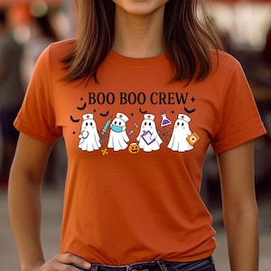 May include: An orange t-shirt with the text "BOO BOO CREW" and a graphic of five cartoon ghosts, each with a different Halloween-themed accessory. The woman is wearing blue jeans and is standing outside.