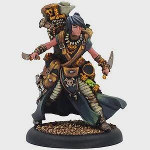 Może przedstawiać: Ręcznie malowana miniaturowa figurka postaci fantasy, pozująca z dwoma sztyletami. Postać ma na sobie szczegółowy strój z plecakiem, sakwami i powłóczystym płaszczem. Paleta kolorów obejmuje odcienie brązu, zieleni i złota, z czarną podstawą.
