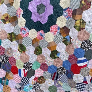 May include: A patchwork quilt with a central floral motif. The quilt showcases a large, six-petal flower in purple and green, encircled by a ring of hexagonal fabric pieces in diverse colours and patterns. The quilt is made from a variety of vintage fabrics.