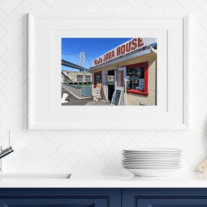 May include: A red and white sign with the text "Red's Java House" above a small brick building with a red door and windows. The building is located on a pier with a bridge in the background.