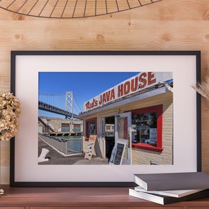 Puede incluir: Un letrero rojo y blanco con el texto "Red's Java House" sobre un pequeño edificio amarillo con bordes rojos. El edificio tiene una ventana grande con un letrero de Budweiser y una ventana más pequeña con un letrero que dice "Champions". El edificio está ubicado en un muelle con un puente azul en el fondo.