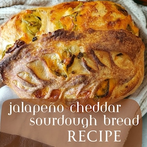 May include: A close-up of a sourdough bread loaf with jalapeno and cheddar cheese. The bread is golden brown and has a crispy crust. The text "jalapeno cheddar sourdough bread RECIPE" is written on a brown background.