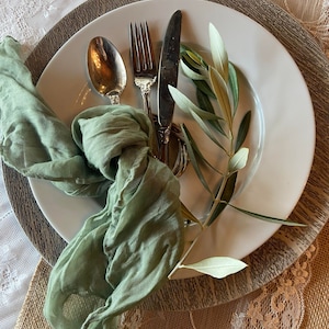 May include: A white plate with silverware, a green cloth napkin, and olive branches. The silverware includes a spoon, fork, and knife. The napkin is knotted and draped on the plate. The olive branches add a natural touch to the table setting.