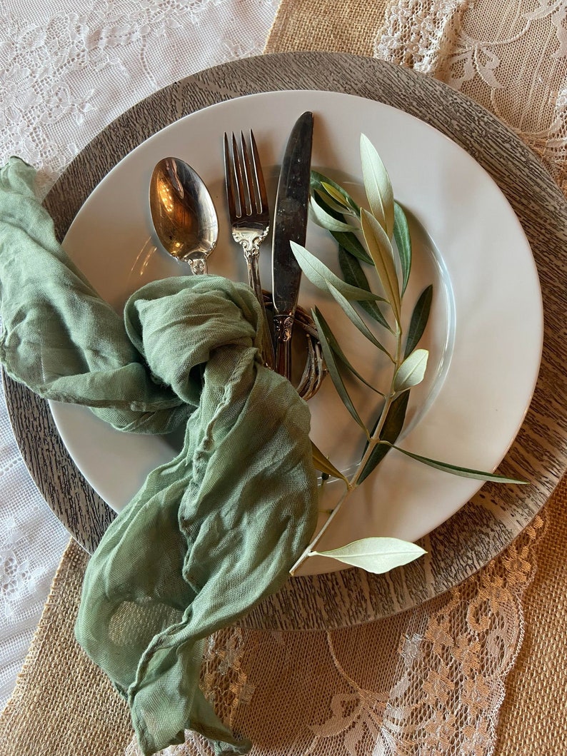 May include: A white plate with silverware, a green cloth napkin, and olive branches. The silverware includes a spoon, fork, and knife. The napkin is knotted and draped on the plate. The olive branches add a natural touch to the table setting.