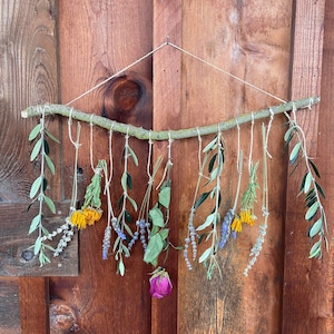 May include: A rustic wall hanging featuring a natural branch adorned with various dried flowers and greenery. The arrangement includes lavender, yellow flowers, and a pink rose, suspended by twine against a wooden background.