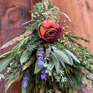 May include: A dried floral arrangement featuring a deep red rose as the focal point, surrounded by green herbs, lavender, and wheat. The bouquet is tied with twine, creating a rustic and natural aesthetic.