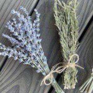 May include: Two bundles of dried herbs on a gray wooden surface. One bundle features lavender with purple flowers, and the other has green herbs. Both are tied with twine.