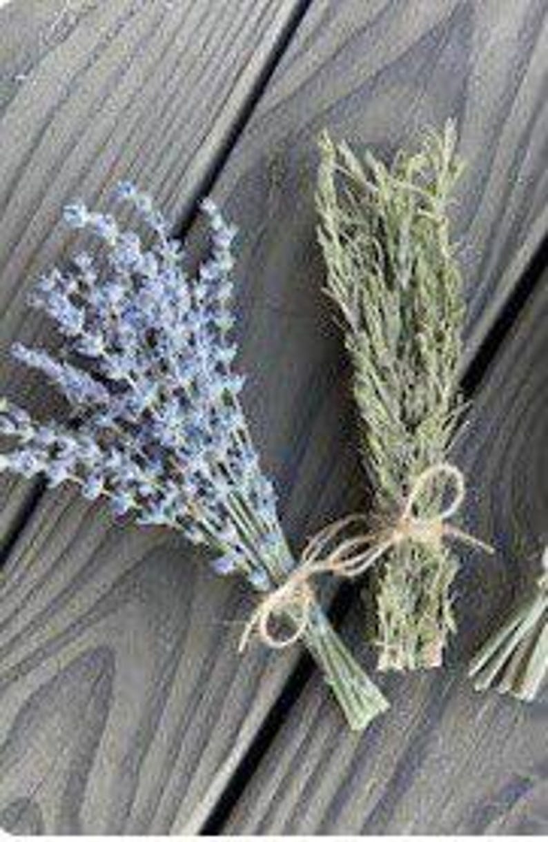 May include: Two bundles of dried herbs on a gray wooden surface. One bundle features lavender with purple flowers, and the other has green herbs. Both are tied with twine.