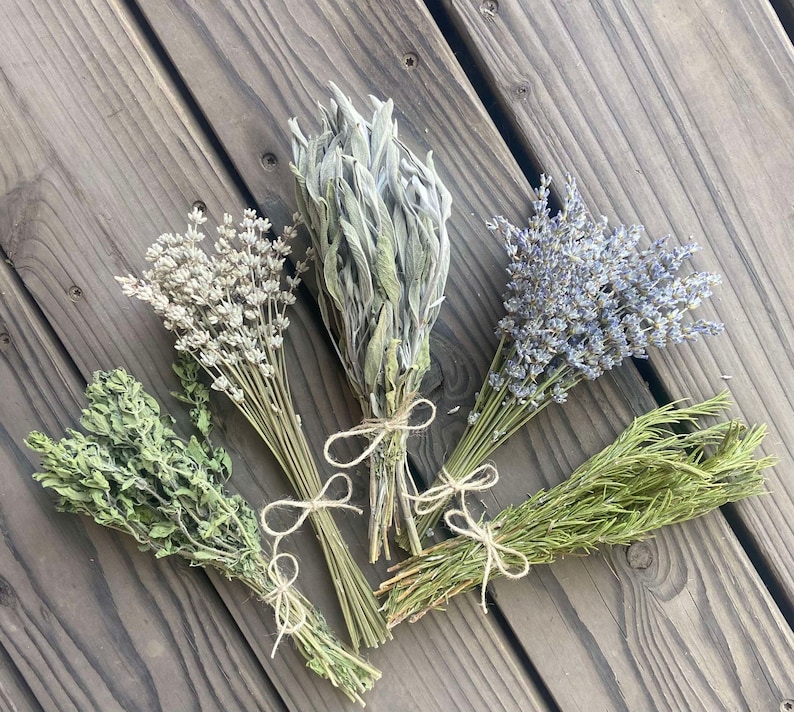 May include: Five bundles of dried herbs, including lavender, sage, and rosemary, tied with twine. The herbs are various shades of green and purple, arranged on a weathered wooden surface.