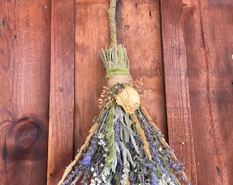 May include: A decorative broom made from a natural tree branch handle and a bundle of dried flowers and herbs. The arrangement includes purple lavender, white baby's breath, green sage leaves, golden wheat stalks, and a single dried cream-colored rose.