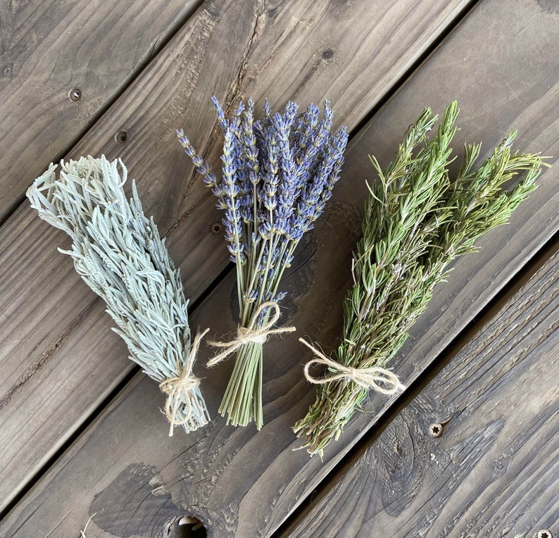 May include: Three bundles of dried herbs tied with twine. The bundle on the left is a pale green color, the center bundle is purple, and the bundle on the right is green. The bundles are arranged on a dark wooden surface.