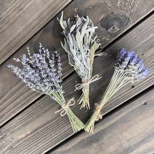 May include: Three bundles of dried herbs tied with twine on a wooden surface. The bundles include lavender with purple flowers, sage with silvery-green leaves, and another lavender bundle. The wooden surface has a dark brown color.