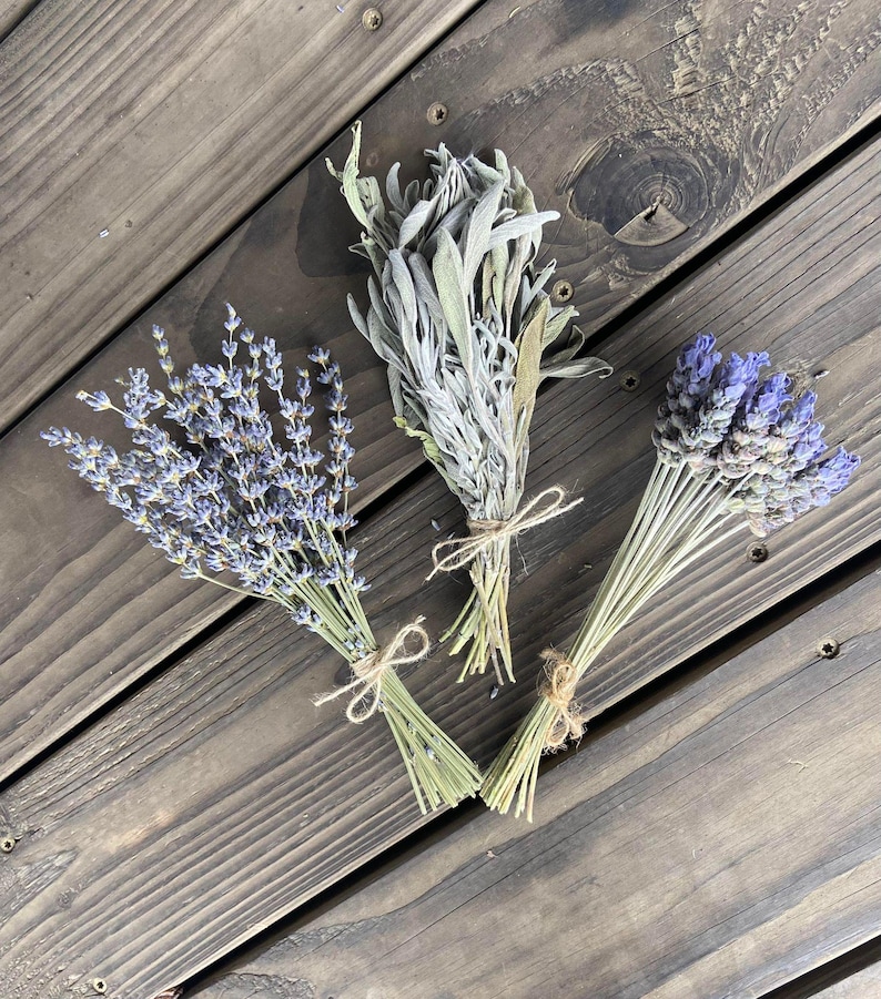 May include: Three bundles of dried herbs tied with twine on a wooden surface. The bundles include lavender with purple flowers, sage with silvery-green leaves, and another lavender bundle. The wooden surface has a dark brown color.