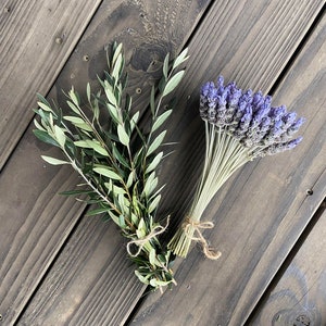May include: Two bundles of dried botanicals on a wooden surface. One bundle features green olive branches tied with twine. The other bundle showcases purple lavender, also tied with twine. The wood has a dark, weathered appearance.
