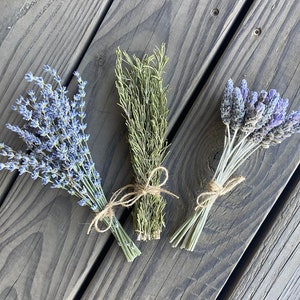 May include: Three bundles of dried herbs tied with twine. The bundles include lavender with purple flowers, rosemary with green needles, and lavender with purple flowers. The herbs are arranged on a weathered wooden surface.